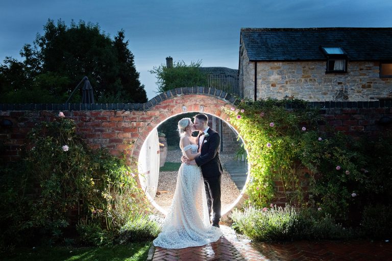 Bride and groom kiss together whilst being lit up by a light through a moonstone circular brick doorway. Elegant photo by Blooming Photography. Taken at Blackwell Grange, Warwickshire.