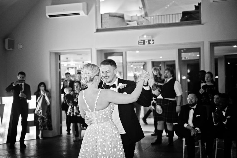 First dance candid photo by Blooming Photography. Taken at Blackwell Grange, Warwickshire. B&w image of a bride and groom doing their first dance, guests look onwards behind them