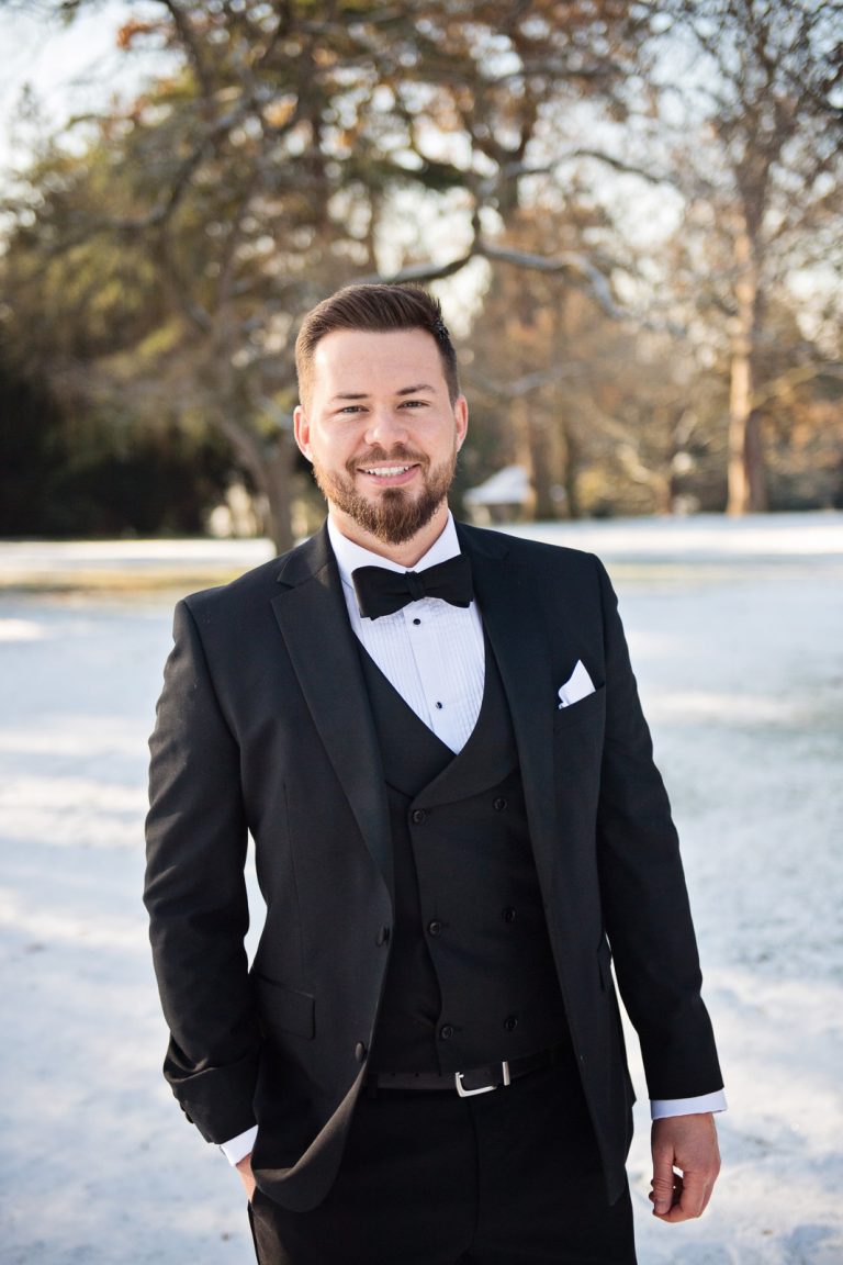 A smart groom stands fashionably with one hand in his pocket, with the winter snow behind him. Photograph by Ben Roberts Blooming Photography, taken at Eastington Park.