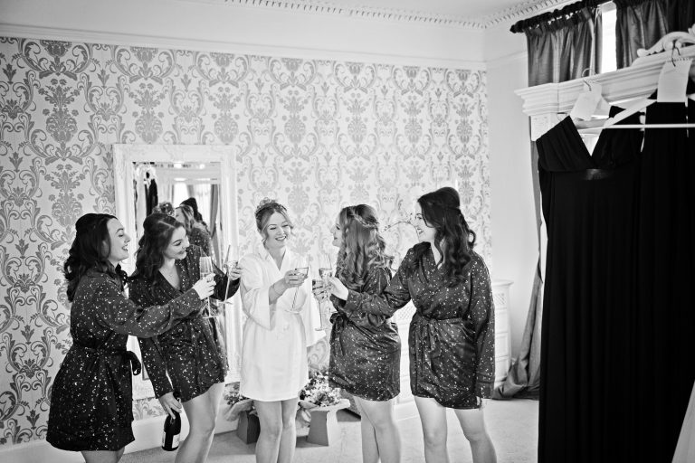 B&W photograph of brides in their wedding gowns cheering to each other with a glass of fizz. Photographed at Eastington Park Winter Wedding by Blooming Photography