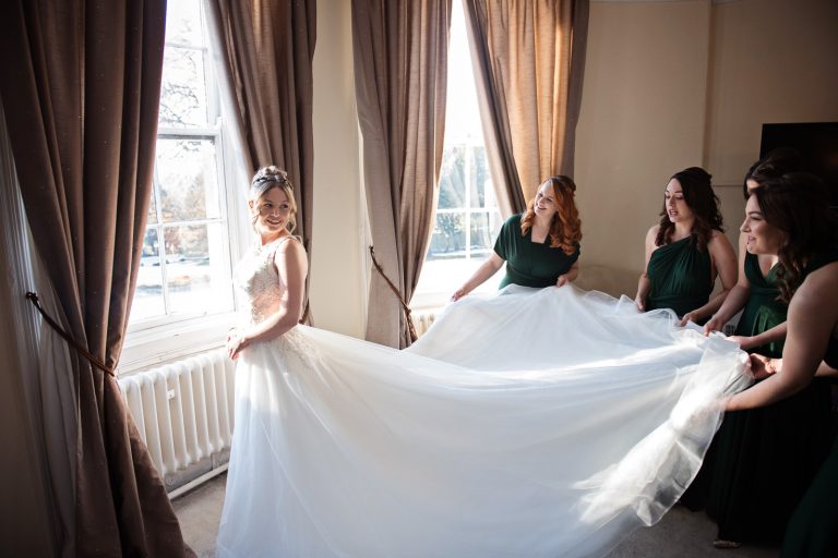 Bridesmaids hold up the brides wedding dress in front of the bay window at Eastington Park. Tajen by Blooming Photography