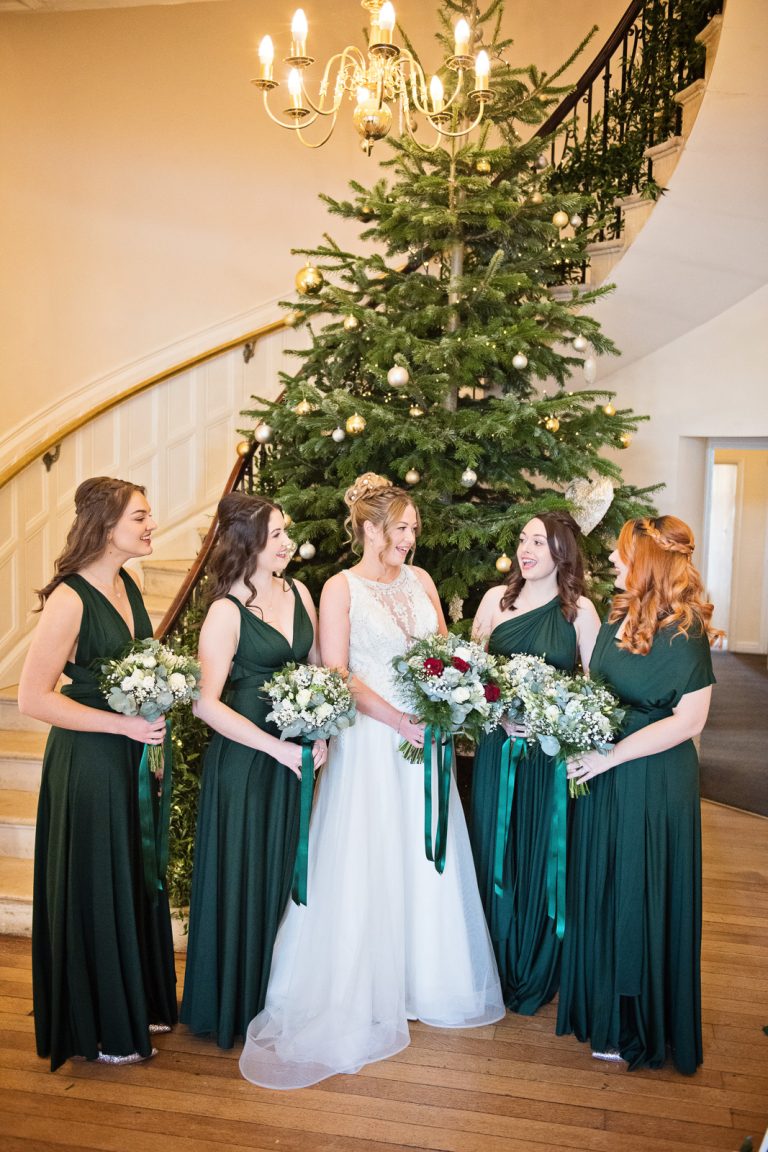 Bride and her bridesmaids stand together at the bottom of the majestic stairs at Eastington Park, with a Christmas tree behind them.