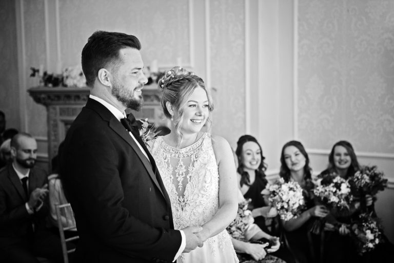 B&W photo of Bride and groom smiling towards the registra's (not seen), with the bridesmaids smiling behind them.