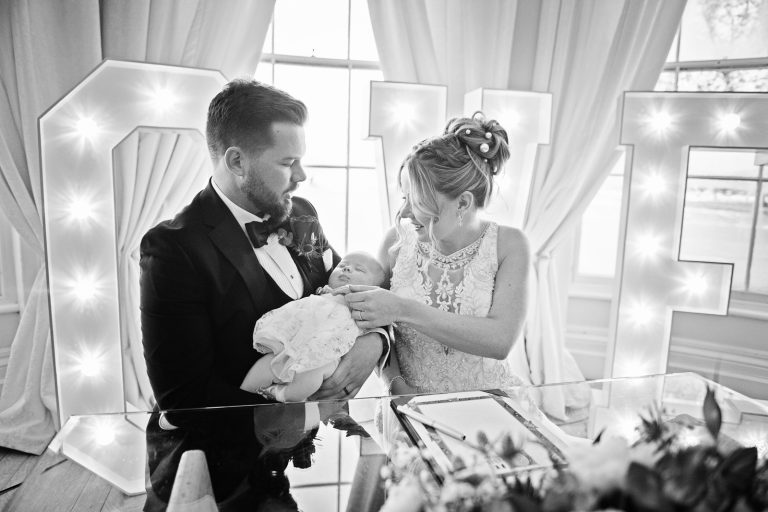 B&W image of Bride and groom hold their baby in front of the bay windows at Eastington Park, with large lit up Love letters behind them.