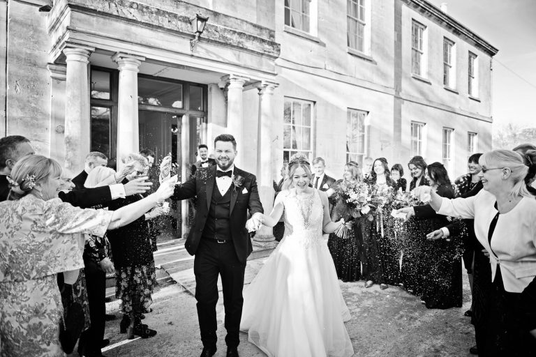 B&W photo of bride and groom being showered with confetti by their wedding guests outside the front of Gloucestershire wedding venue, Eastington Park