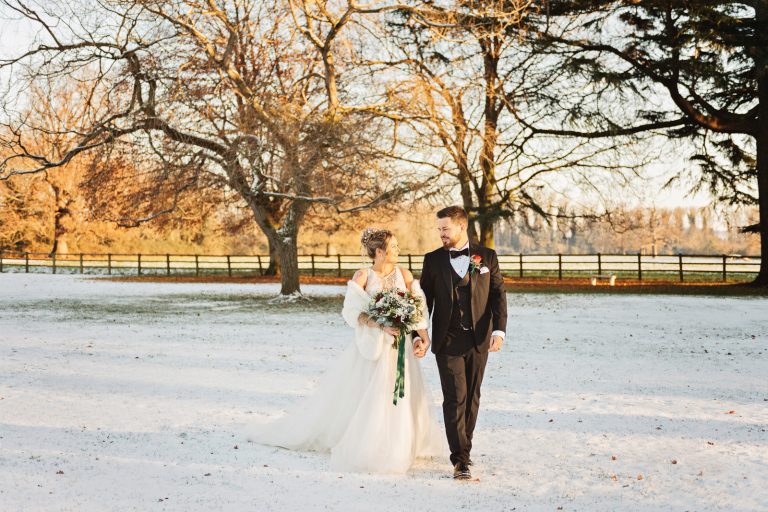 Bride and groom walking hand in hand together in the snowy grounds of Eastington Park. Photo by Blooming Photography