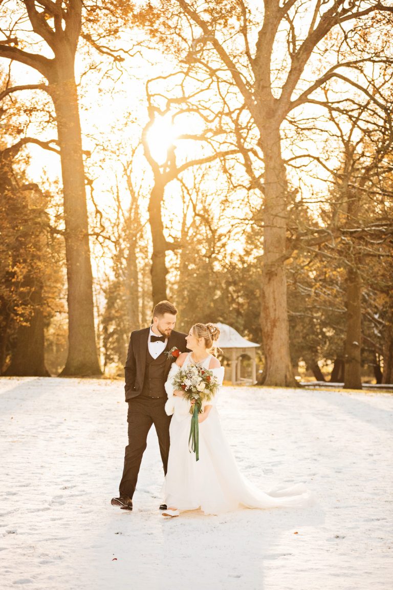 Majestic portrait photo of bride and groom walking together across snow, with large mature trees behind them in the golden sun behind them. Photo by Blooming Photography