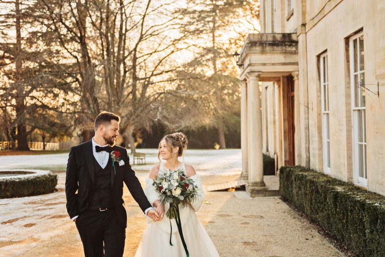 Bride & Groom hold hands whilst smiling at each other as they walk in the snowy grounds of Eastington Park.