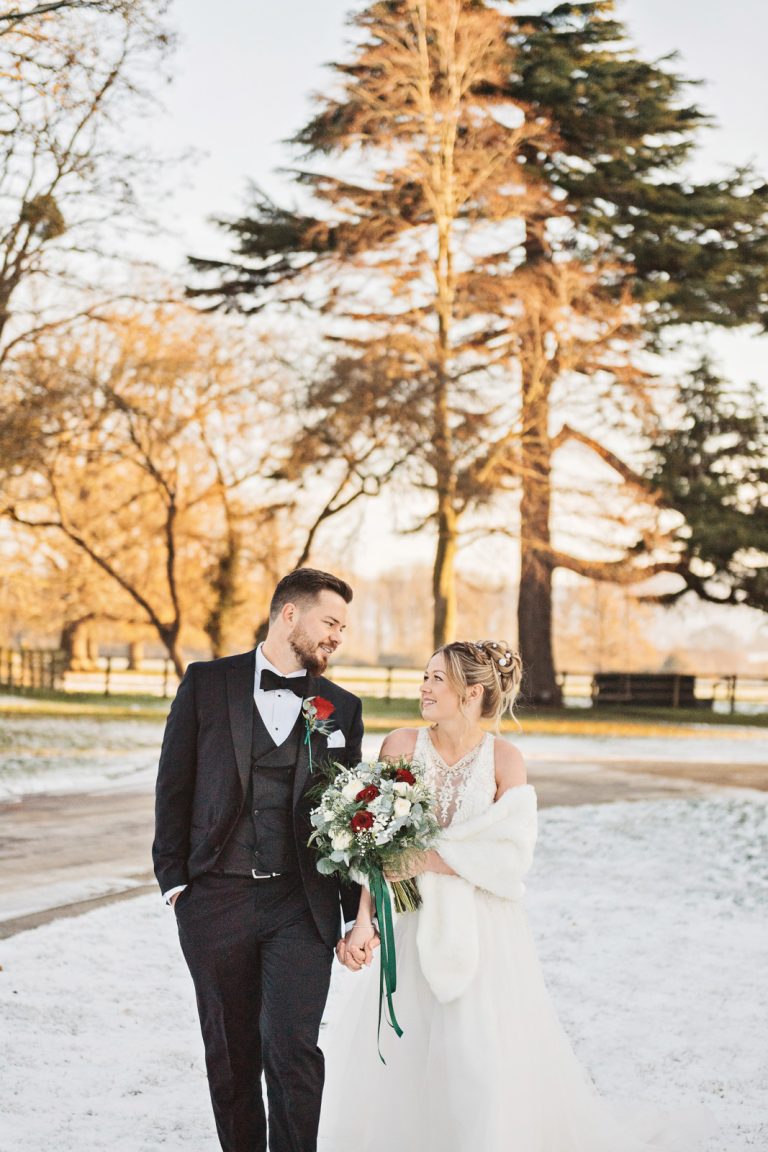 Portrait photo of Bride & Groom holding hands whilst smiling at each other as they walk in the snowy grounds of Eastington Park.