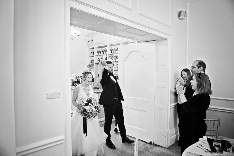 B&W candid photo of bride and groom entering their wedding breakfast room with their guests cheering.