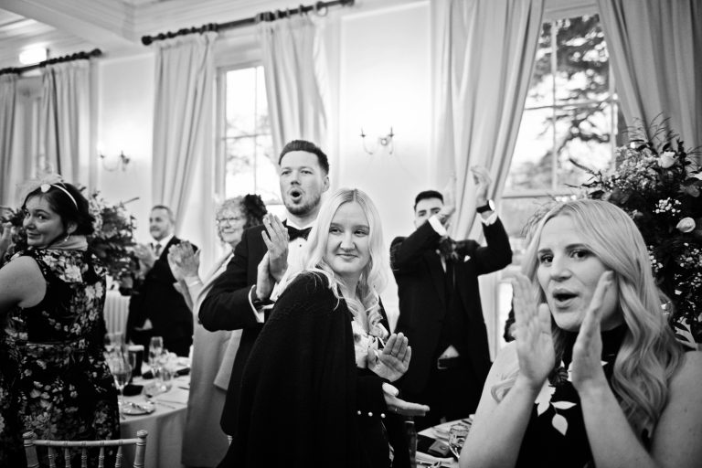 B&W photo of bride and grooms friends and family cheering as the bride and groom enter the wedding breakfast room.