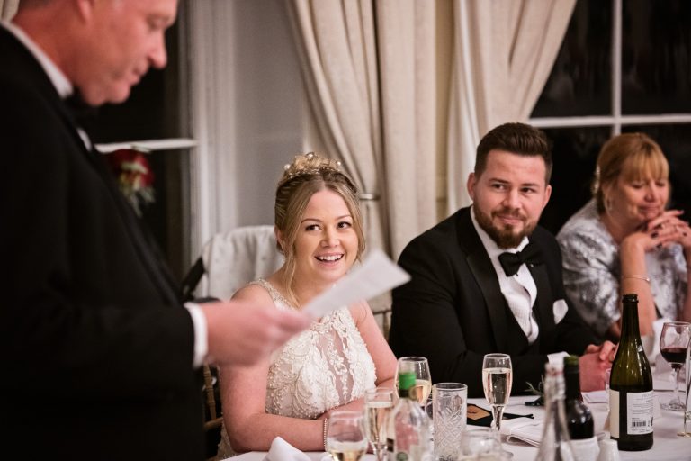 The bride and groom on the wedding top table smile and laugh at the father of the brides speech. Photograph by Blooming Photography taken at Eastington Park at Christmas Wedding.