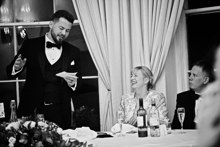 delivers his wedding speech in front of the wedding guests. Grooms Mum is laughing. Black and white photograph by Blooming Photography taken at Eastington Park at Christmas Wedding.