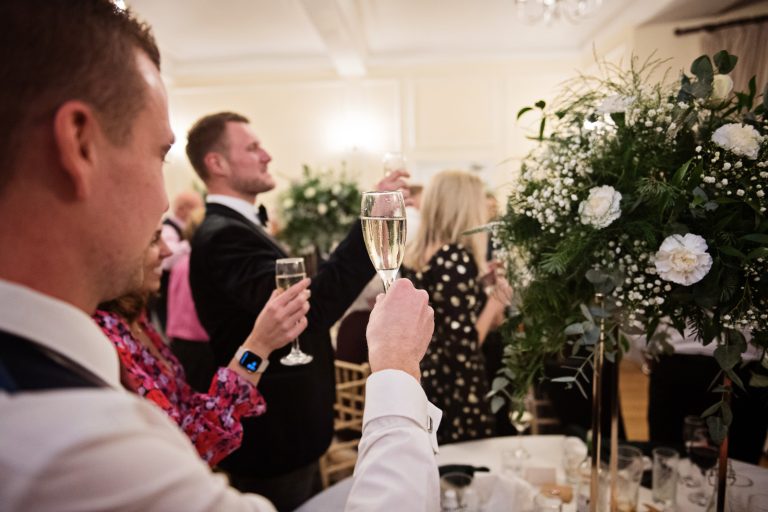 Guest raises their champagne glasses to toast the end of the speeches. A floral bouquet in the mid right of the picture. Photograph by Blooming Photography taken at Eastington Park at Christmas Wedding.