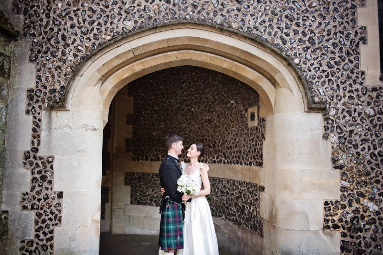 Natural moments photo of bride and groom holding each other under medieval arch Winchester, photo by Blooming Photography.