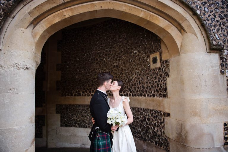 Natural wedding photo of bride and groom kissing under medieval arch Winchester, photo by Blooming Photography.