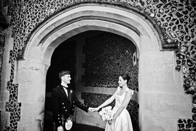 Candid black & white wedding photo of bride and groom holding hands medieval arch Winchester, photo by Blooming Photography.