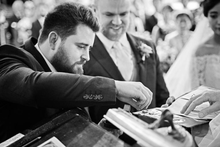 The best man places the rings onto a mat watched by the bride and groom and vicar. Story telling black and white photograph by Blooming Photography.