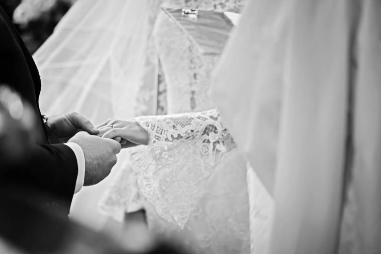 A groom places a wedding ring on his brides finger. Close up story telling black and white photograph, by Blooming Photography.