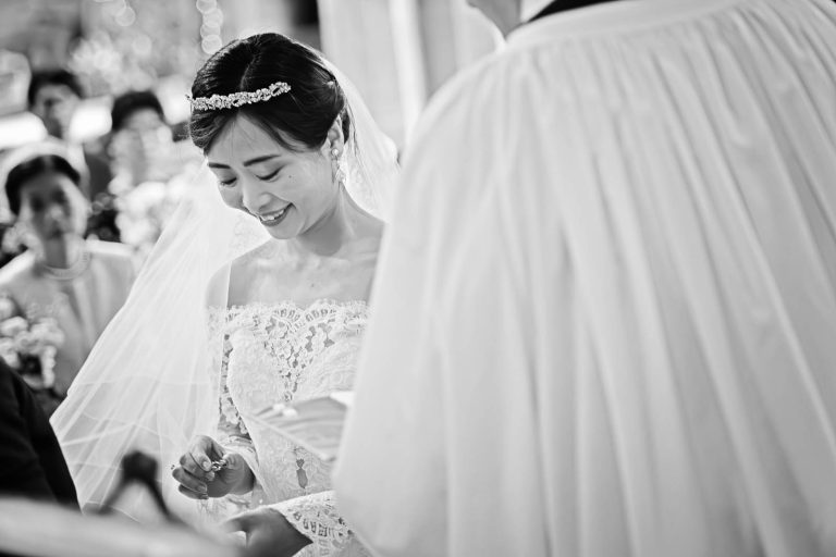 A bride smiling as she is about to place a wedding ring on her husband to be's finger. Story telling black and white photograph by Blooming Photography.
