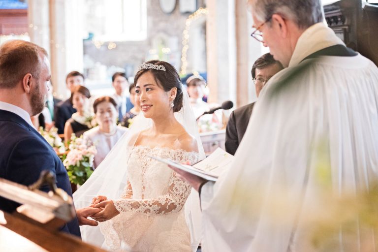A bride smiling as she is about to place a wedding ring on her husband to be's finger. Story telling photograph by Blooming Photography.