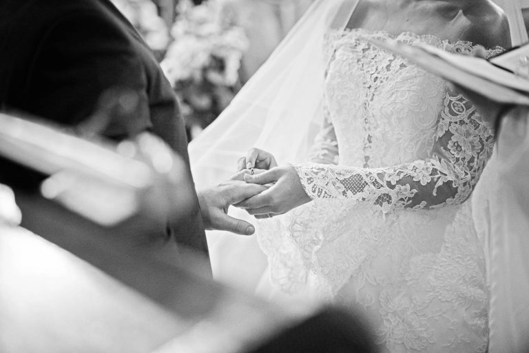A close up of bride placing a wedding ring on her husband to be's finger. Story telling black and white photograph by Blooming Photography.