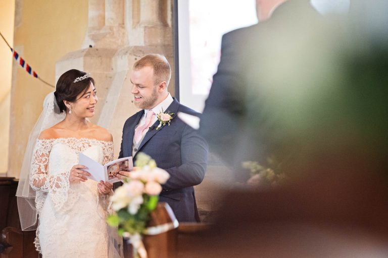 A bride and groom smiling at each other whilst they sing a hymn during the wedding ceremony. Story telling photograph by Blooming Photography.