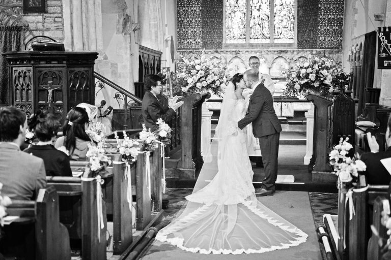Bride and groom kiss as they are announced husband and wife. Bride and groom are central to the image, wooden wedding pews filled with guests (who are clapping in celebration). Story telling black and white photograph by Blooming Photography.