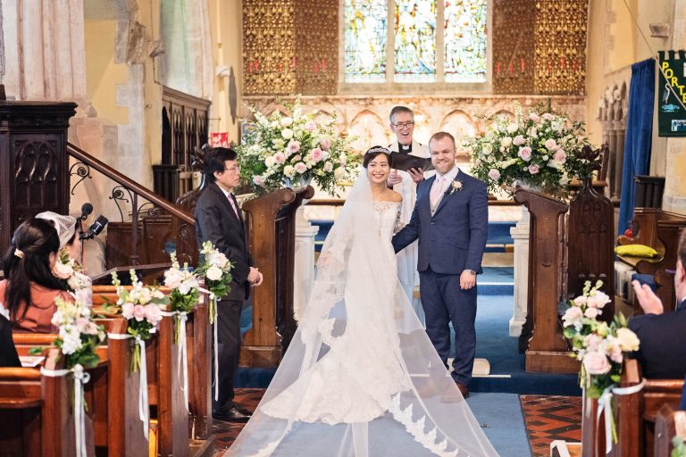 Bride and groom smile as they are announced husband and wife. Bride and groom are central to the image, wooden wedding pews filled with guests (who are clapping in celebration). Story telling photograph by Blooming Photography.