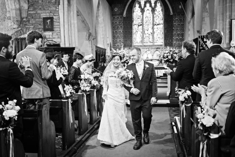 Bride and groom walk down the isle smiling with their wedding guests clapping. Story telling black and white photograph by Blooming Photography.