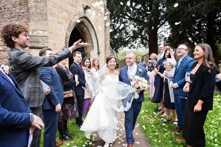 Story telling photograph of bride and groom exiting the church whilst being showered with confetti. Photograph by Blooming Photography.