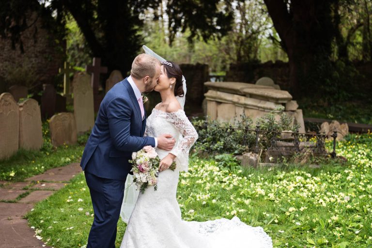 A bride and groom kiss in the grounds of the church. Cowslips flowering in the grass and graves in the background. Candid photograph by Blooming Photography.