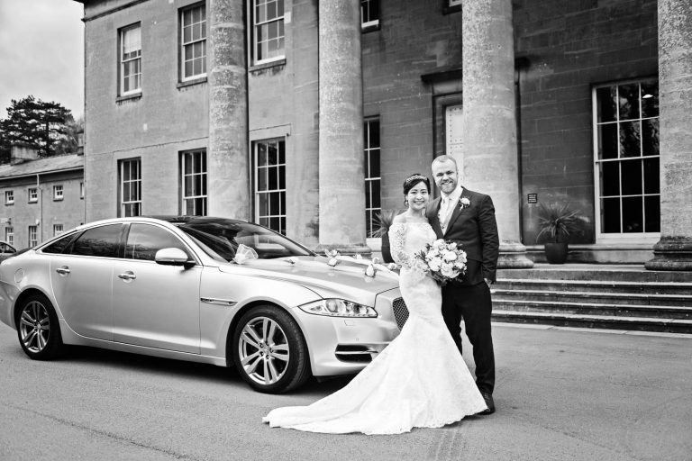 A bride and groom stand smiling and facing the camera, next to a Jaguar wedding car and outside grand Leigh Court entrance. Black and white photo by Blooming Photography.