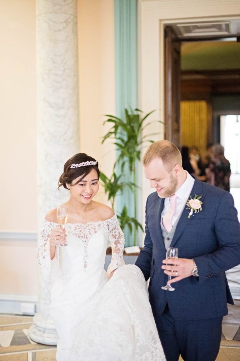 Portrait photograph of bride and groom hold champagne glasses in the entrance hall of Leigh Court. Elegant photograph by Blooming Photography.