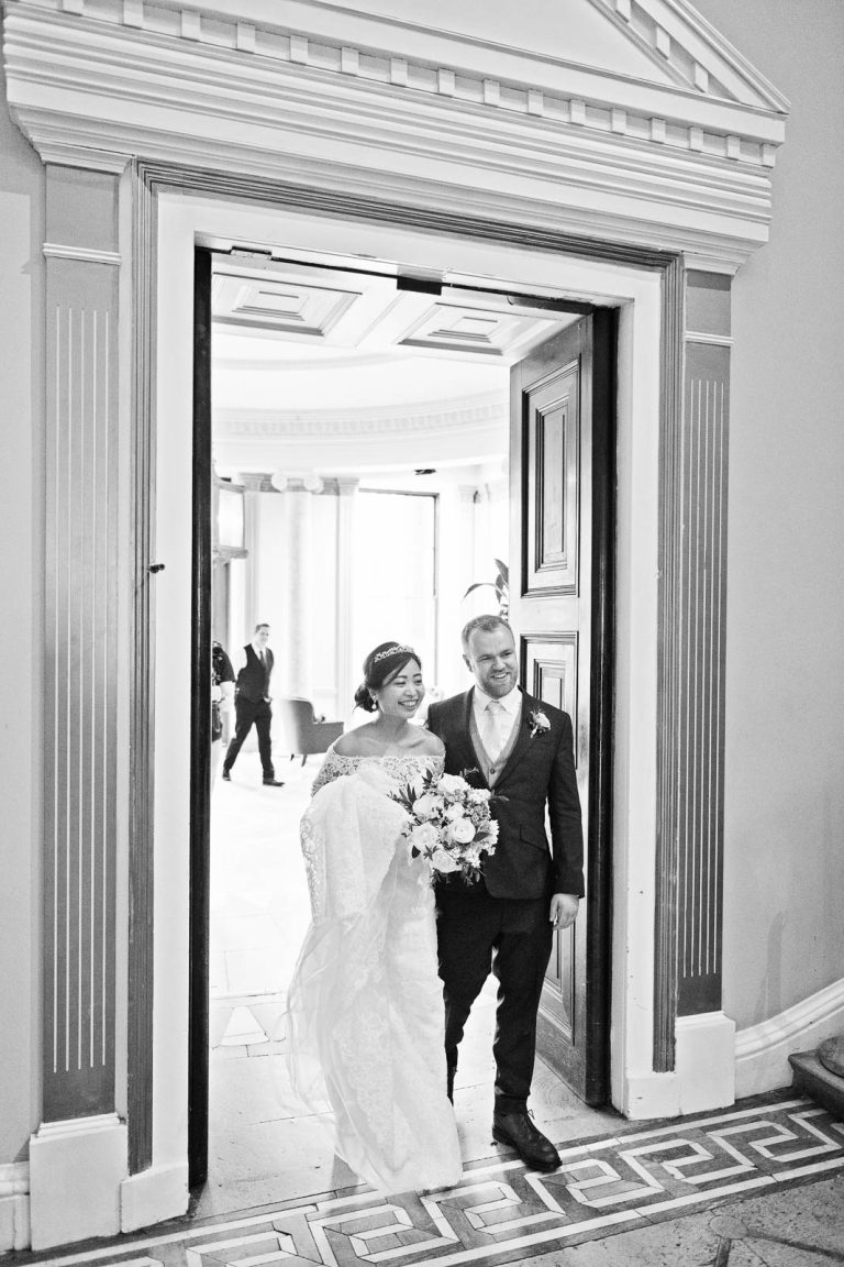 Bride and groom enter through a grand door at Leigh Court. Candid black and white photograph by Blooming Photography.