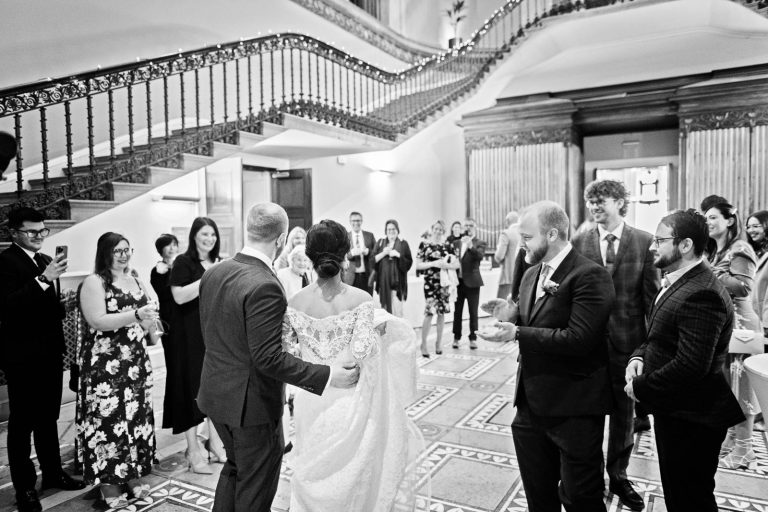 Bride and groom enter Leigh Court, their wedding guests clap and cheer their arrival. Candid black and white photograph by Blooming Photography.