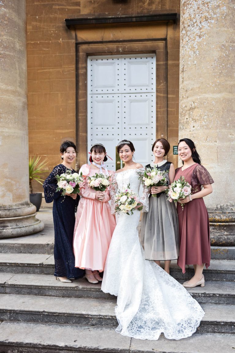 Formal photograph of bride and bridesmaids stand on the steps of Leigh Court, Bristol, holding their wedding flowers. Candid photograph by Blooming Photography.