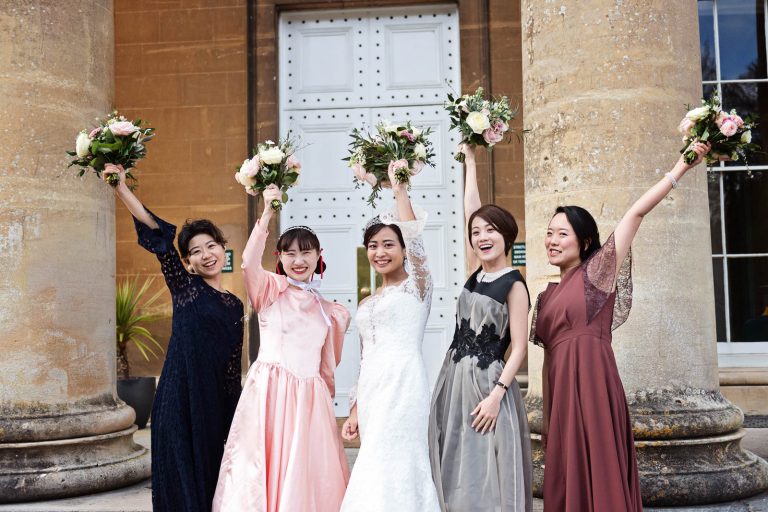 Bride and bridesmaids stand on the steps of Leigh Court, Bristol, holding their wedding flowers up in the air. Candid photograph by Blooming Photography.
