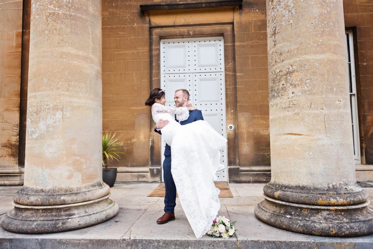Groom lifts his bride up into his arms in the middle of the stone pillars at Leigh Court, Bristol. Bridal bouquet on the floor. andid photograph by Blooming Photography.