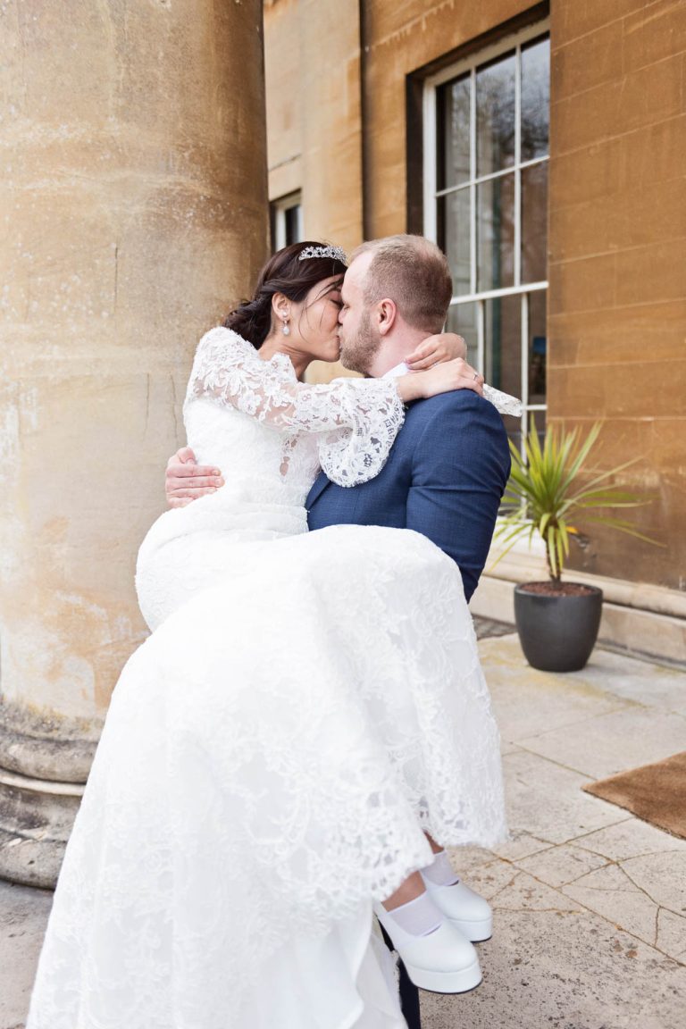 Portrait photo of the Groom lifting his bride up into his arms in the middle of the stone pillars at Leigh Court, Bristol. Candid photograph by Blooming Photography.