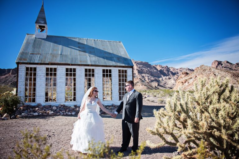 Bride and groom hold hands amongst cacti and to the side of a wedding chapel in the Nevada desert at Nelson Ghost Town.