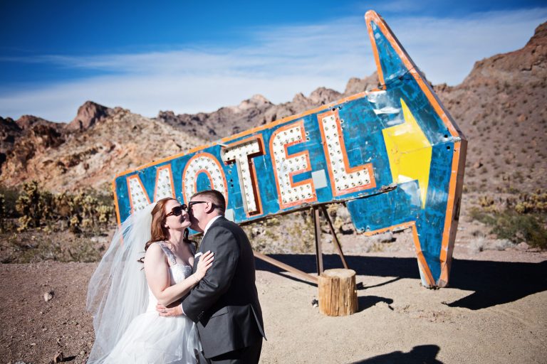 A bride and groom stand in front of a very large old Las Vegas Motel and Arrow sign in the Nevada Desert. The groom is kissing the bride on the cheek. Photograph by Blooming Photography, photographed at Nelson Ghost Town.