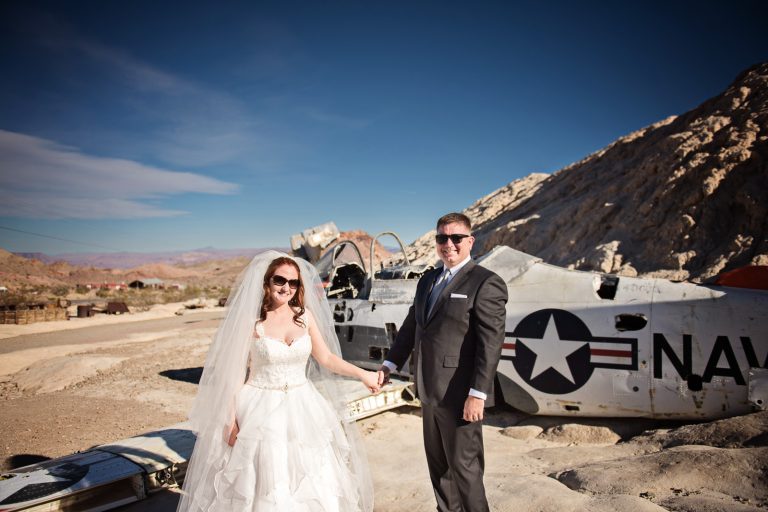 Bride and groom hold hands in the Nevada Desert next to an old US Navy Fighter Plane. Photograph by Blooming Photography, photographed at Nelson Ghost Town.