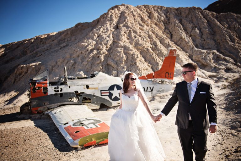 Bride and groom walk and hold hands next to an old US Navy Fighter Plane. Looks cool. Photograph by Blooming Photography, photographed at Nelson Ghost Town.