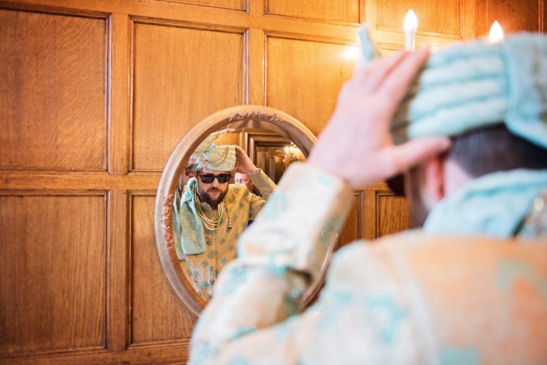 Groom putting on his Indian headpiece in the mirror at North Mymms Park, candidly photographed by Blooming Photography