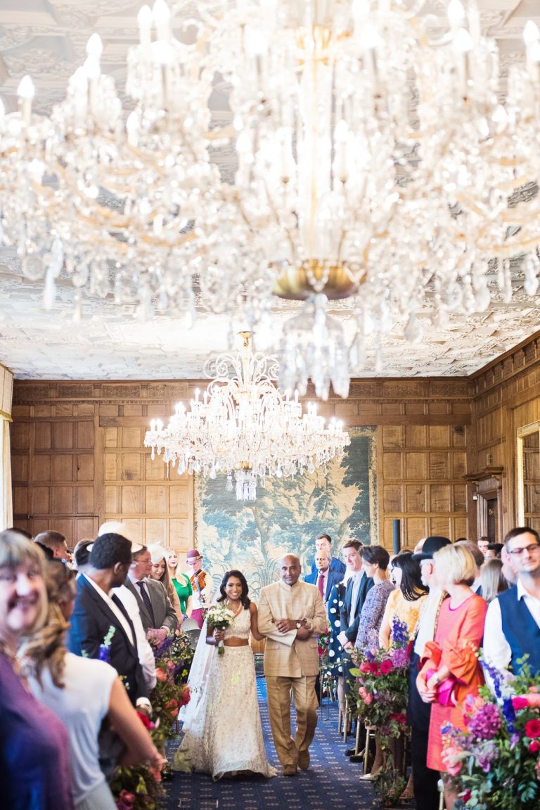 A bride and her father walk down the isle at North Mymms Park. A large chandelier takes up much of the photo giving the photo an elegant look. Candidly photographed by Blooming Photography.