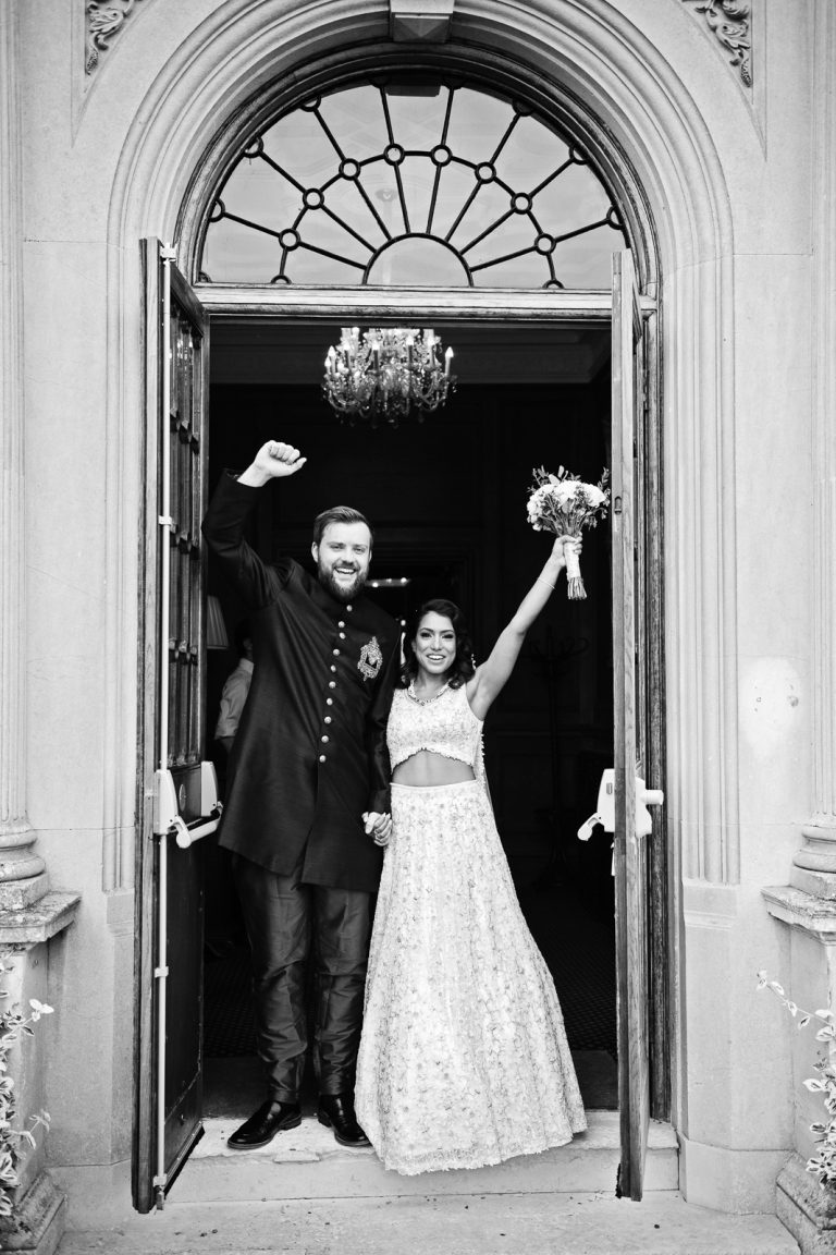 Bride and groom smiling with their hands punching the air as they stand underneath an ornate archway. Black and white image. Photographed at North Mymms Park, by Blooming Photography.)