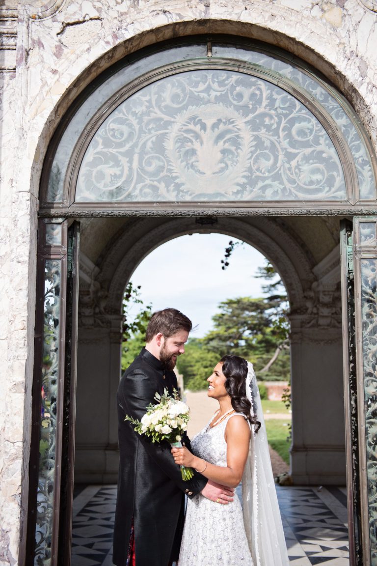 Bride and groom smiling as they stand (tummy to tummy) underneath an ornate archway. Photographed at North Mymms Park, by Blooming Photography.