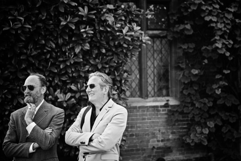 Two gents casually look onward at the wedding party (out of frame), black and white photo. Photographed at North Mymms Park, by Blooming Photography.