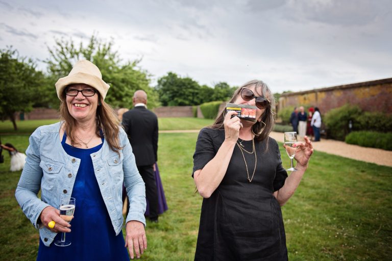 Two wedding guests smile as they are smoking. The one guest is holding up a tobacco pouch up to her mouth. The tobacco pouch has ugly teeth on it. Funny. Photographed at North Mymms Park, by Blooming Photography.